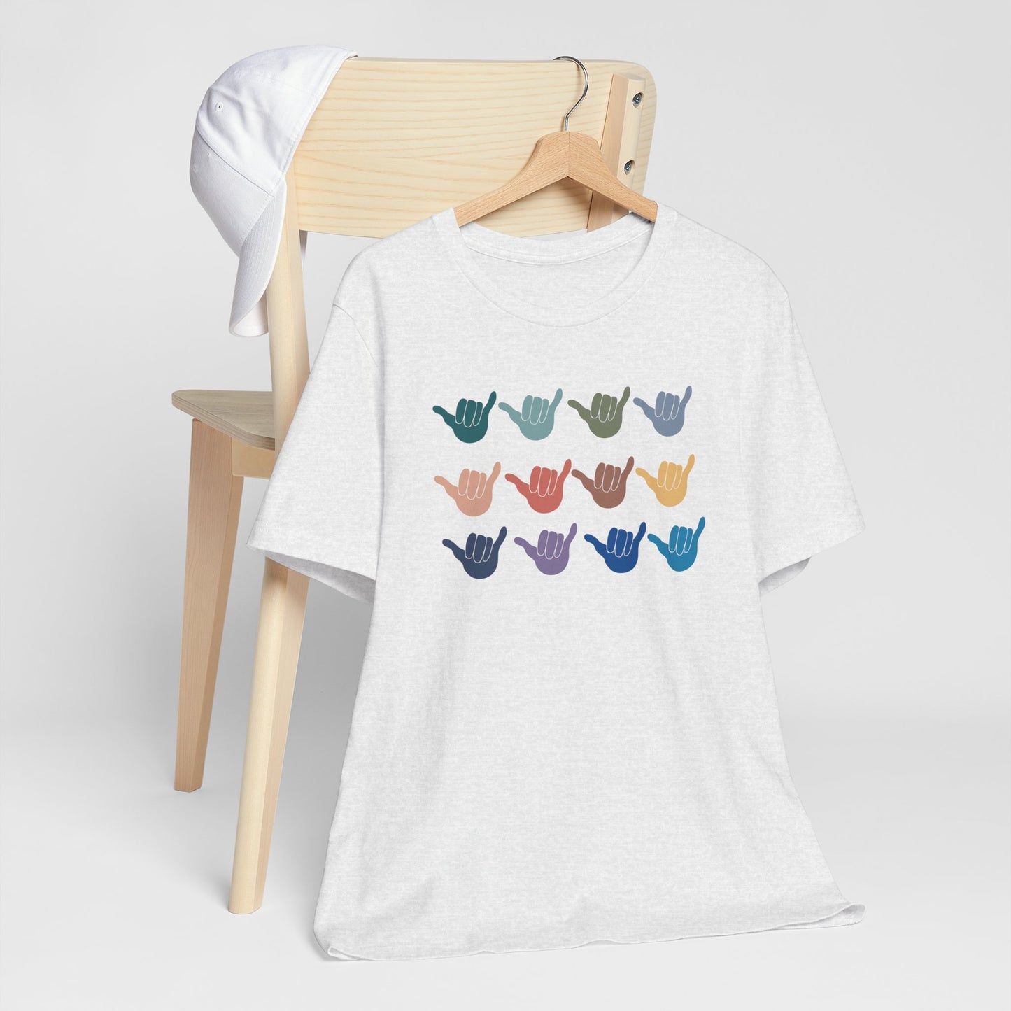 A white heather  short-sleeve t-shirt hanging on a wooden hanger, displayed in front of a light wood chair with a white cap. The t-shirt features a graphic of twelve colorful shaka hand signs arranged in a grid on the chest.