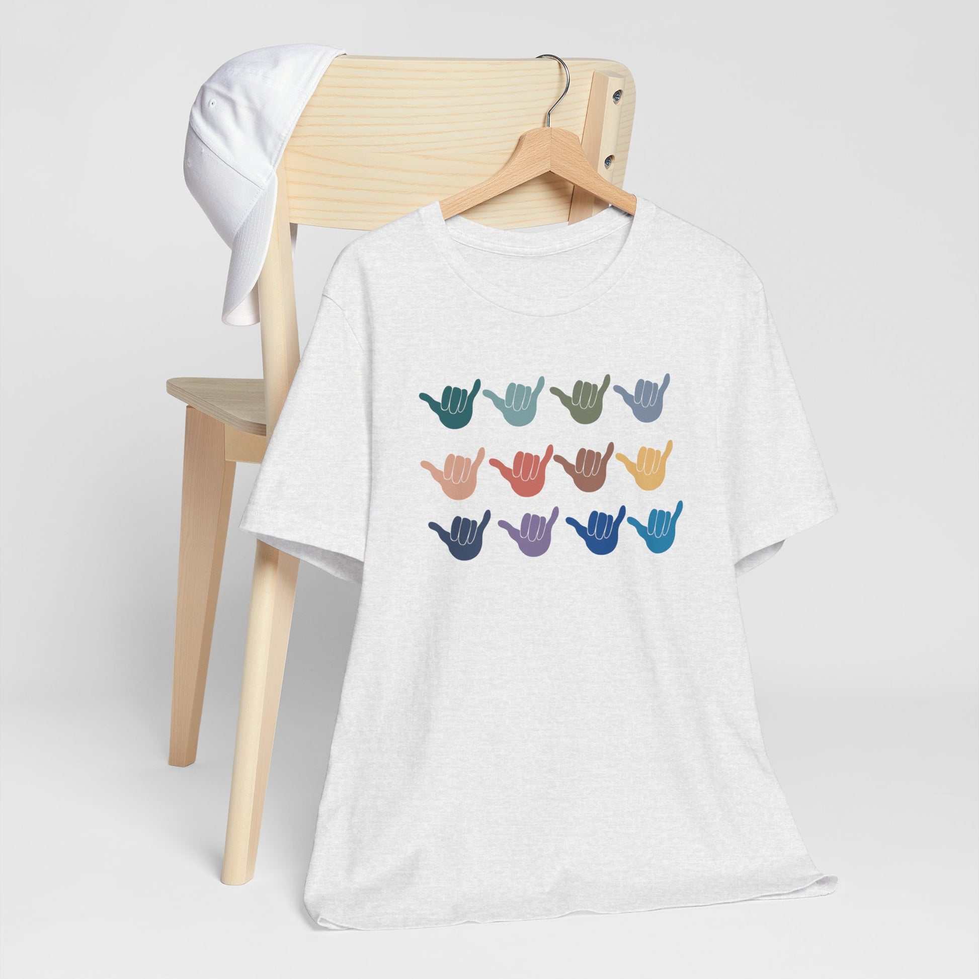 A white heather  short-sleeve t-shirt hanging on a wooden hanger, displayed in front of a light wood chair with a white cap. The t-shirt features a graphic of twelve colorful shaka hand signs arranged in a grid on the chest.