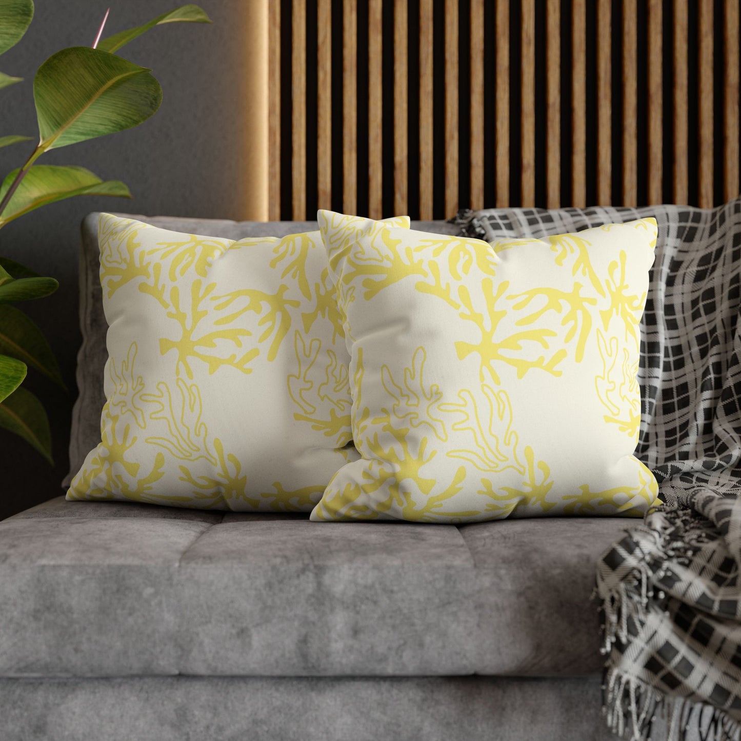 Two square throw pillows with a yellow coral-like botanical pattern on an off-white background, styled on a gray sofa with a black-and-white plaid throw blanket and a wooden slat wall behind them.