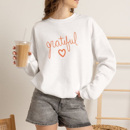 A woman wearing a white crewneck sweatshirt with a graphic that reads 'Grateful' in orange lettering, paired with gray denim shorts and holding an iced coffee.