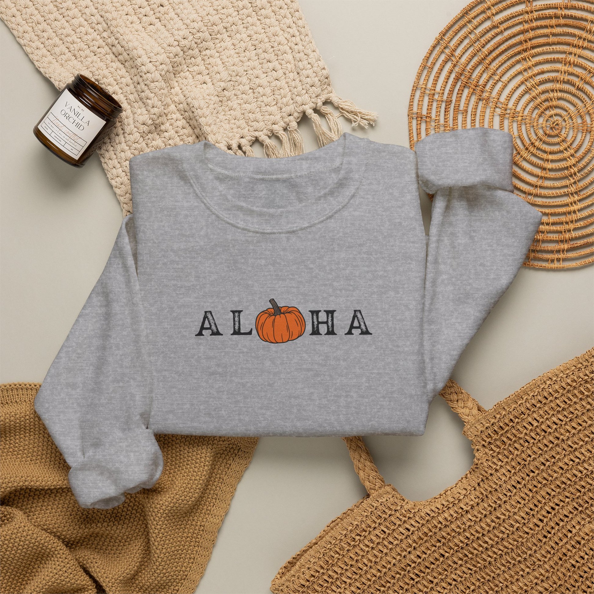 A folded heather gray sweatshirt with 'ALOHA' printed across the chest, where the O is replaced by an orange pumpkin graphic. The sweatshirt is styled in a flat lay with a woven beige blanket, a small amber candle, a round woven placemat, and a tan tote bag on a light neutral background.