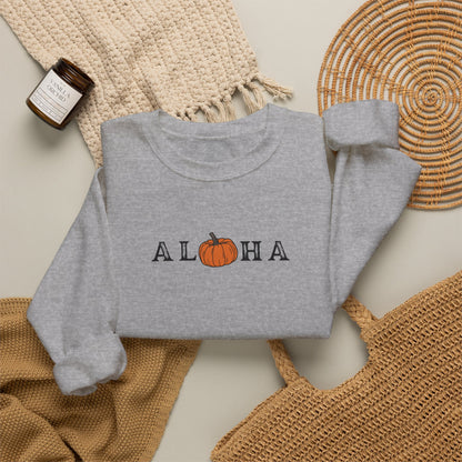 A folded heather gray sweatshirt with 'ALOHA' printed across the chest, where the O is replaced by an orange pumpkin graphic. The sweatshirt is styled in a flat lay with a woven beige blanket, a small amber candle, a round woven placemat, and a tan tote bag on a light neutral background.