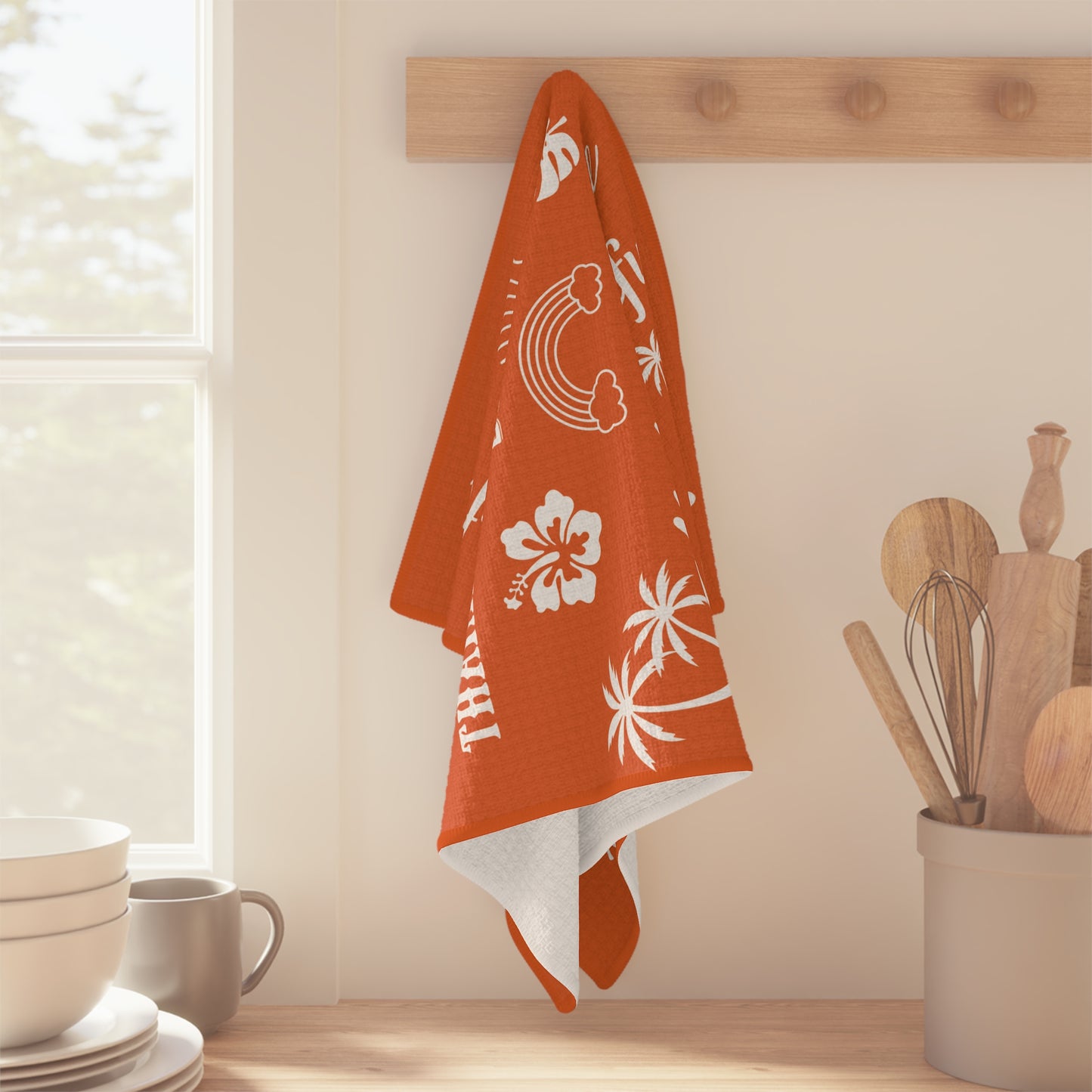An orange kitchen towel with white tropical and Hawaiian-themed prints hangs from a wooden peg on a wall in a bright kitchen setting.