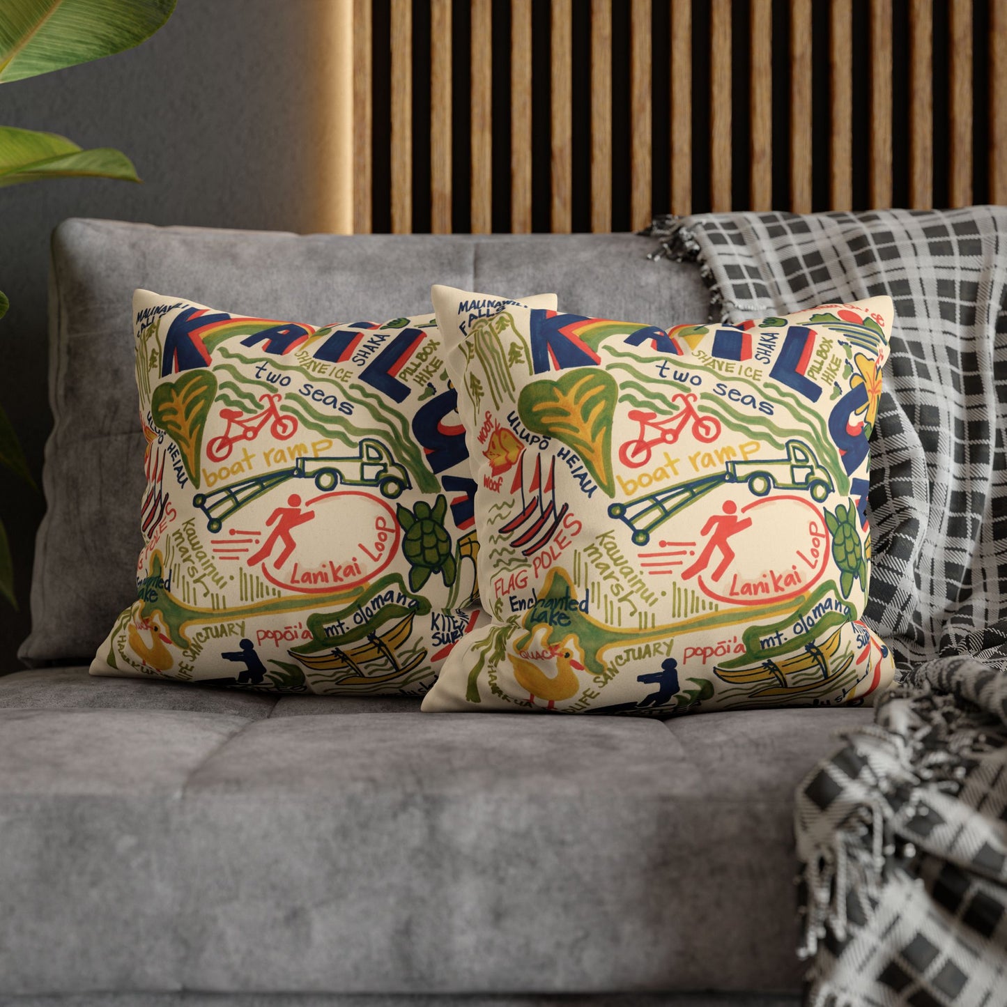 Two square throw pillows with a colorful vintage-style illustrated print featuring bicycles, people, and text of Kailua, Oahu styled on a gray sofa with a plaid throw blanket.