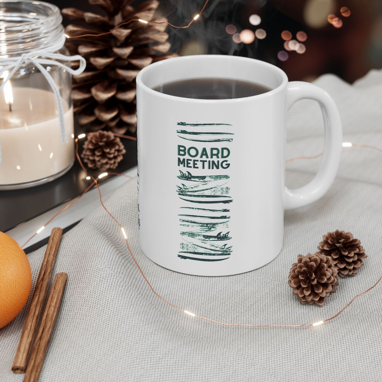 A white ceramic mug filled with a dark beverage sits on a light fabric surface, surrounded by holiday decor including pinecones, string lights, cinnamon sticks, an orange, and a lit candle. The mug has a green printed design with the words 'BOARD MEETING' and brushstroke-style lines.