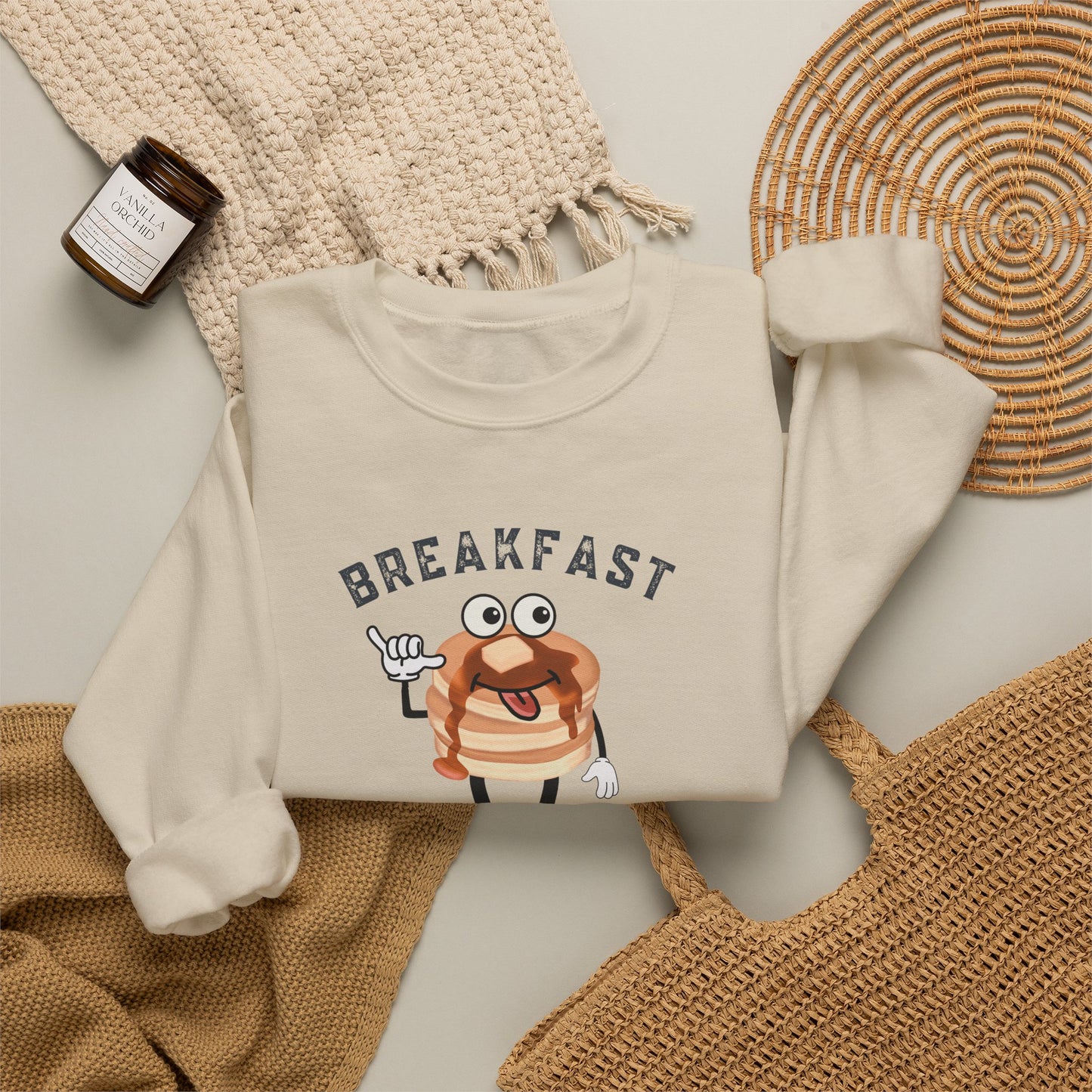 A folded cream sweatshirt with a cartoon pancake graphic and the word 'BREAKFAST' printed across the chest, styled in a flat lay with a woven blanket, a small candle, and a wicker tray.