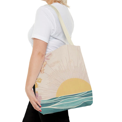 A woman wearing a white t-shirt and black pants carries a canvas tote bag with a retro sunset and ocean design. The bag has a cream-colored strap and features a graphic print of a yellow sun rising over teal blue waves with pink floral accents.
