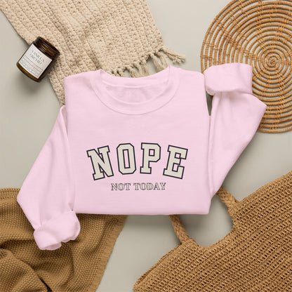 A light pink crewneck sweatshirt with the words 'NOPE NOT TODAY' printed on the front, styled in a flat lay with a beige knit blanket, a woven basket, and a small candle.
