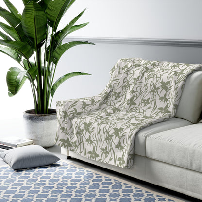 Sage green orchid-and-leaf print sherpa fleece blanket with a warm white background and rounded corners used as a throw blanket on a grey couch.
