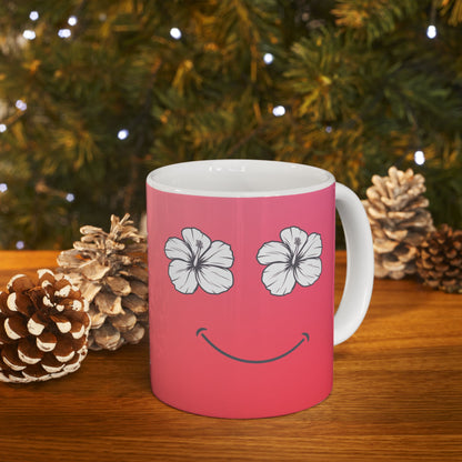 A pink ceramic mug with a white handle and interior, featuring a smiley face design made of two white hibiscus flowers for eyes and a curved smile. The mug sits on a wooden surface with pinecones and blurred Christmas tree branches in the background.