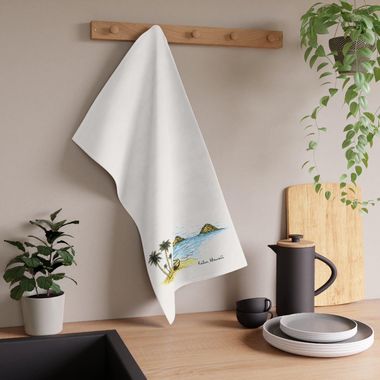 A white kitchen towel with a colorful beach illustration and the text 'Kailua Hawaii” hanging from a wooden rack on a beige wall above a wooden countertop with dishes and plants.