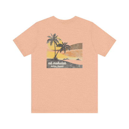A heather peach short-sleeve t-shirt with a vintage-style graphic on the back depicting two palm trees against a yellow and orange sunset, with the text 'ná makulua' and 'Kailua, Hawaii!' printed below.