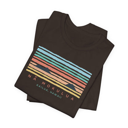 A folded dark brown t-shirt with a colorful retro sunset graphic and the text 'NÁ MOKULUA KAILUA, HAWAII' on the front, displayed on a clean white background.