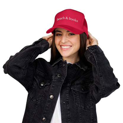 Person wearing a red trucker cap with embroidered text 'beach & books' and a black denim jacket on a white background