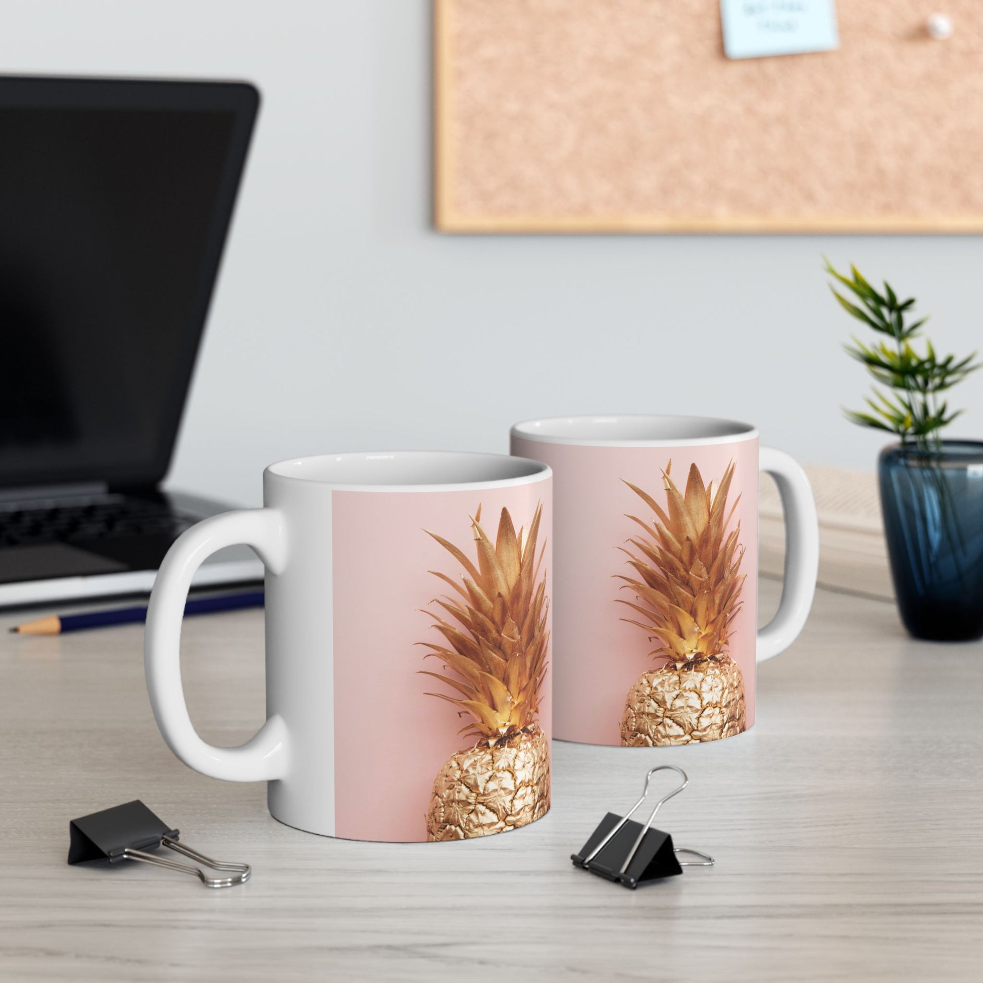 Two white ceramic mugs with a pink and gold pineapple graphic, displayed on a light wood desk with office props like a laptop, binder clips, and a small plant.