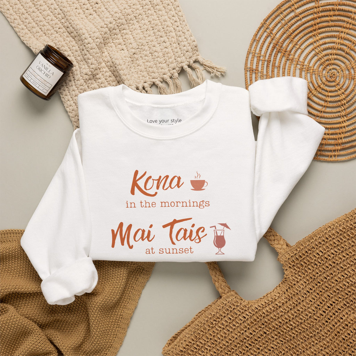 A white crewneck sweatshirt folded neatly on natural accessoires featuring orange text that reads 'Kona in the mornings Mai Tais at sunset' with small coffee cup and cocktail glass illustrations.