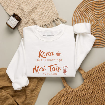 A white crewneck sweatshirt folded neatly on natural accessoires featuring orange text that reads 'Kona in the mornings Mai Tais at sunset' with small coffee cup and cocktail glass illustrations.