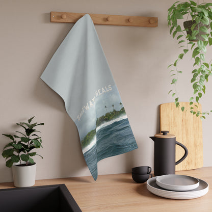 A kitchen towel with a scenic ocean and island print 'saltwater heals' hanging on a wooden wall rack above a kitchen counter with dishes and plants.