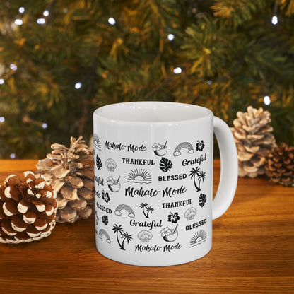 A white ceramic mug with a black tropical-themed print featuring the words 'Mahalo Mode', 'Grateful', and 'Blessed' alongside palm trees, monstera leaves, hibiscus flowers, and sun motifs, displayed on a wooden surface with pinecones and a blurred Christmas tree in the background.