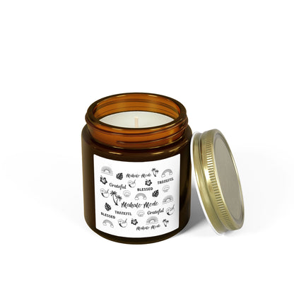 A brown glass jar candle with a gold metal lid and a white paper label featuring black tropical-themed illustrations and text like 'Mahalo Mode,' 'Grateful,' and 'Blessed,' shown on a clean white background.