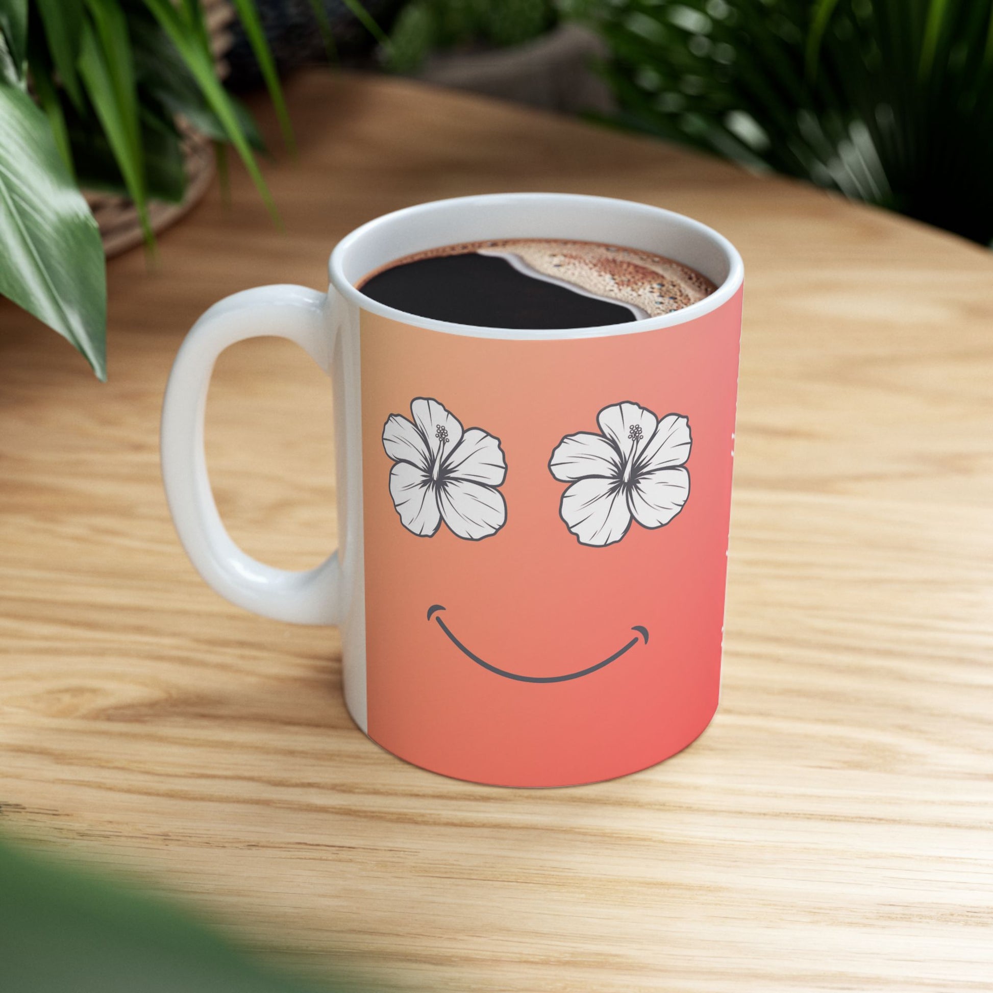 A white ceramic coffee mug with a coral-pink to orange gradient design, featuring a smiley face made of two white hibiscus flowers for eyes and a curved smile. The mug is filled with black coffee and sits on a light wood table with blurred green plants in the background.