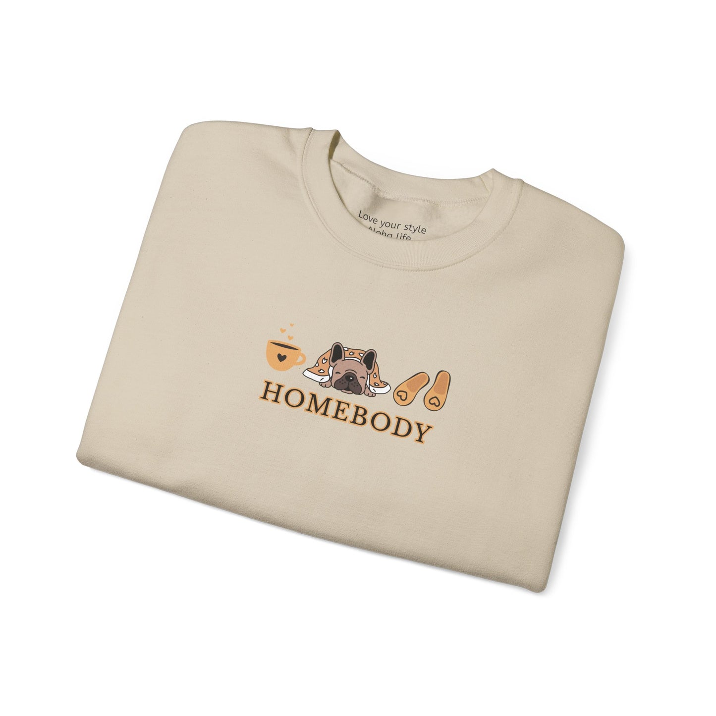 A folded beige crewneck sweatshirt featuring a central graphic of a dog under a blanket, house slippers, coffee cups and the word 'HOMEBODY' printed below it.