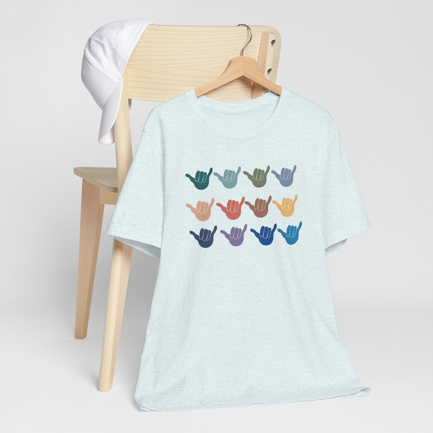 A light heather blue short-sleeve t-shirt hanging on a wooden hanger, displayed in front of a light wood chair with a white cap. The t-shirt features a graphic of twelve colorful shaka hand signs arranged in a grid on the chest.