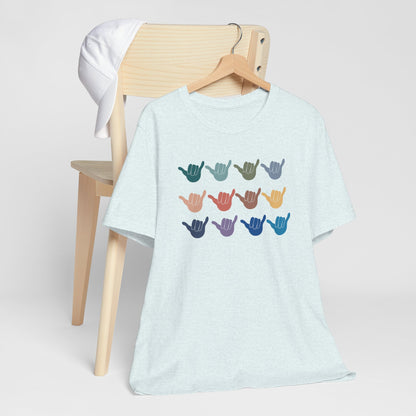 A light heather blue short-sleeve t-shirt hanging on a wooden hanger, displayed in front of a light wood chair with a white cap. The t-shirt features a graphic of twelve colorful shaka hand signs arranged in a grid on the chest.