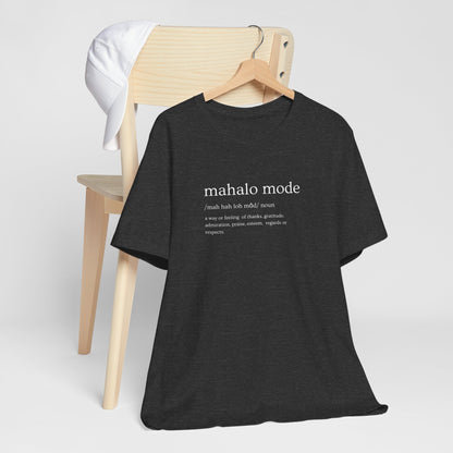 A dark charcoal gray heather t-shirt with a white printed dictionary-style definition of 'mahalo mode' on the chest, hanging from a wooden chair