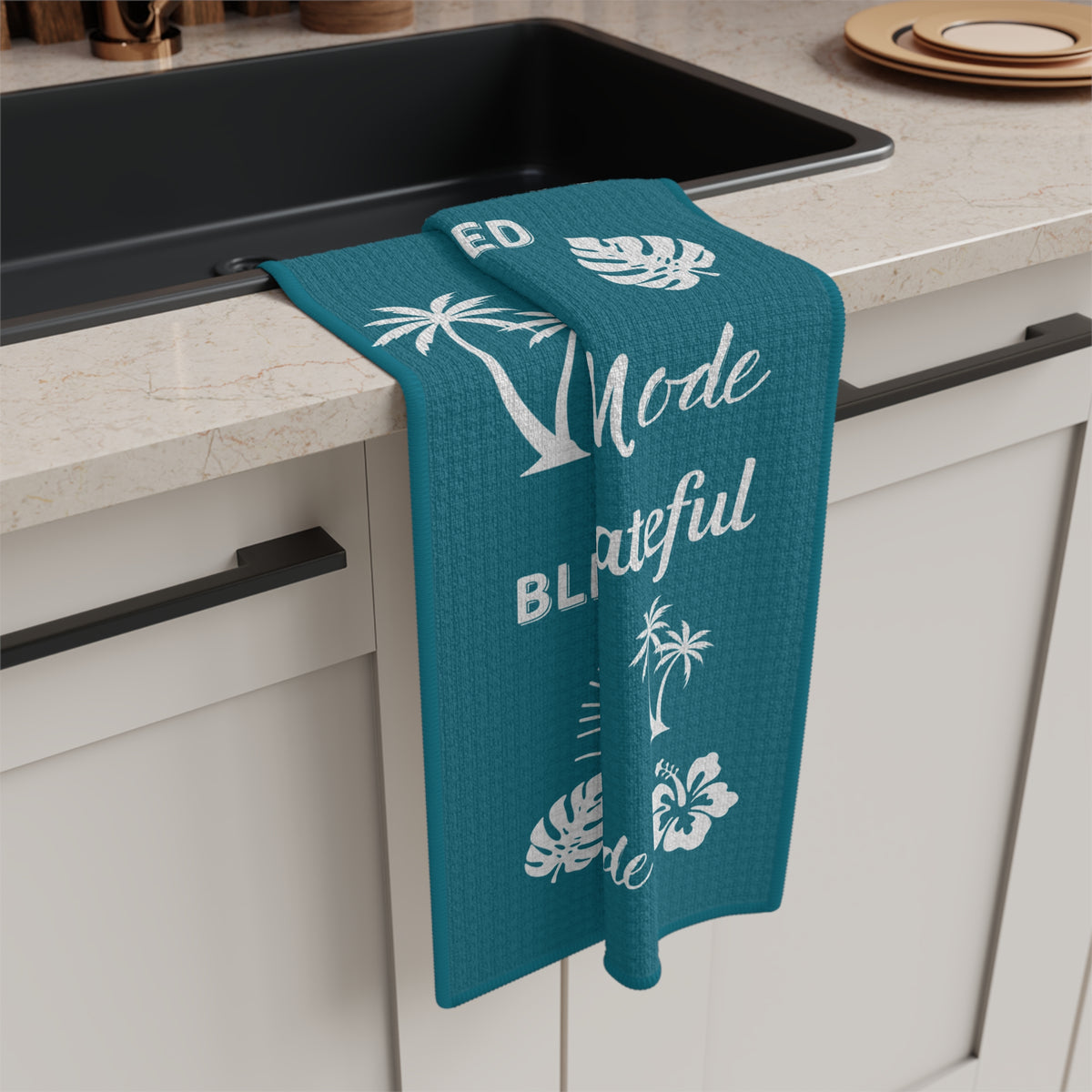 A teal kitchen towel with white tropical and text graphics hangs over the edge of a black sink in a modern kitchen setting.