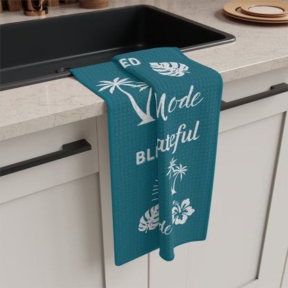 A teal kitchen towel with white tropical and text graphics hangs over the edge of a black sink in a modern kitchen setting.