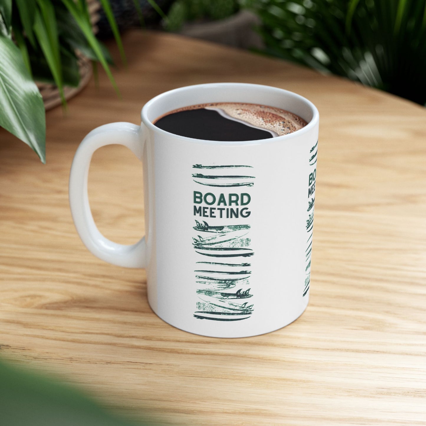 A white ceramic coffee mug with a green 'BOARD MEETING' graphic, filled with black coffee and sitting on a light wood table with blurred green plants in the background.