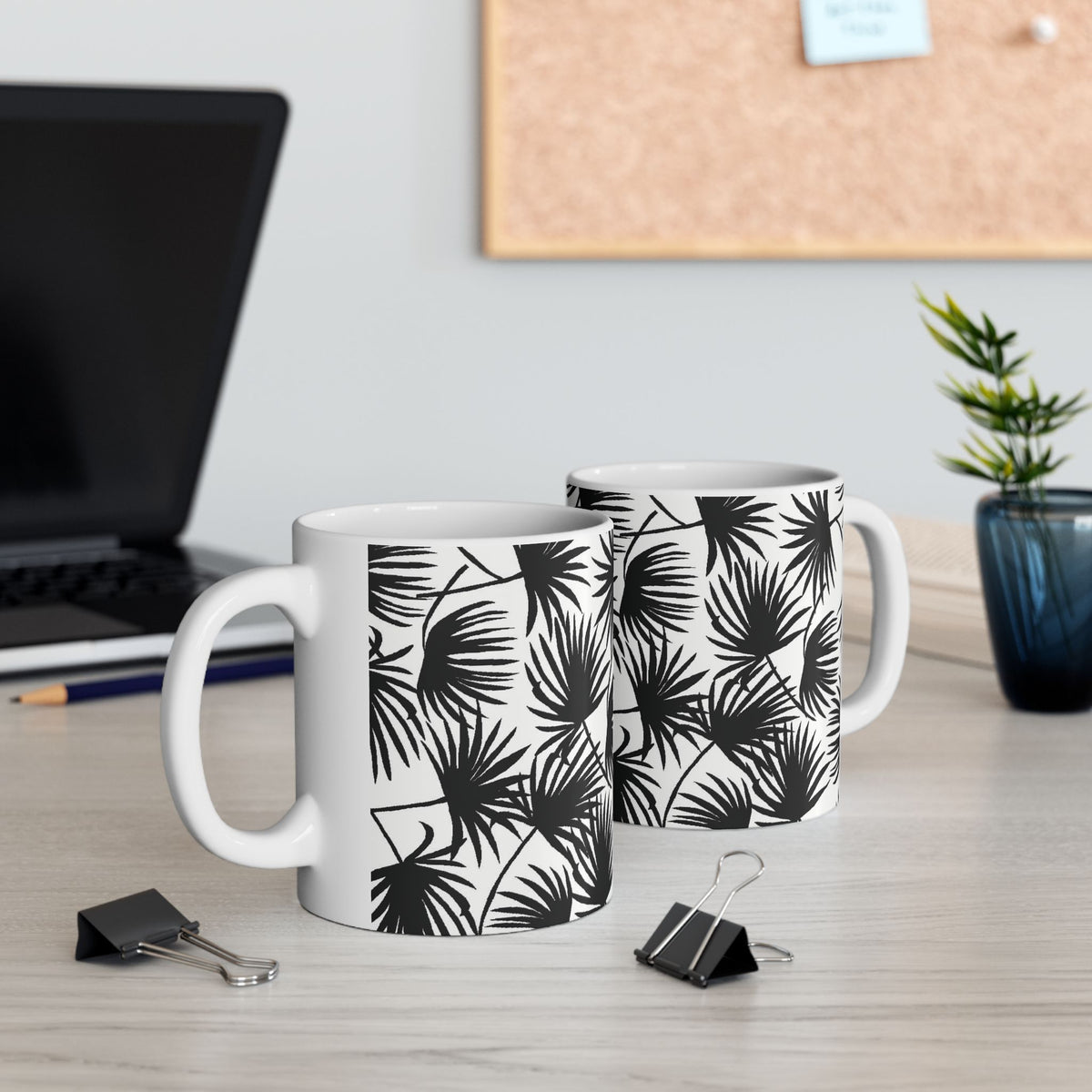 Two Palm Fan Black coffee mugs with black and white tropical palm print by Global Village Kailua sitting on a desk with a computer and binder clips.
