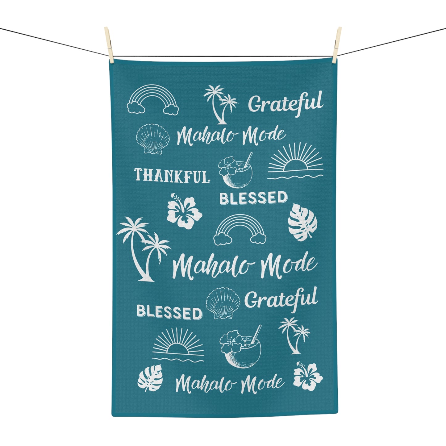 A rectangular teal kitchen towel with white tropical-themed graphics and phrases including 'Mahalo Mode', 'Blessed', 'Grateful', and palm tree, sun, and shell illustrations, hanging from a string with wooden clips against a white background.