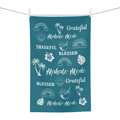 A rectangular teal kitchen towel with white tropical-themed graphics and phrases including 'Mahalo Mode', 'Blessed', 'Grateful', and palm tree, sun, and shell illustrations, hanging from a string with wooden clips against a white background.