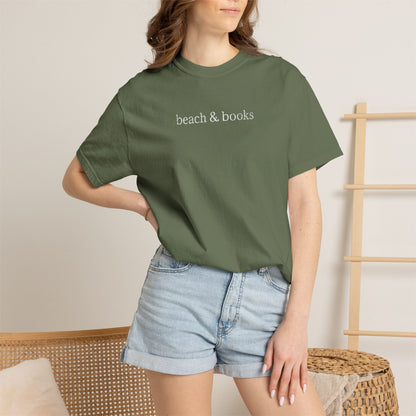 Person wearing a green comfort colors t-shirt with embroidered text 'beach & books' and denim shorts on a neutral background