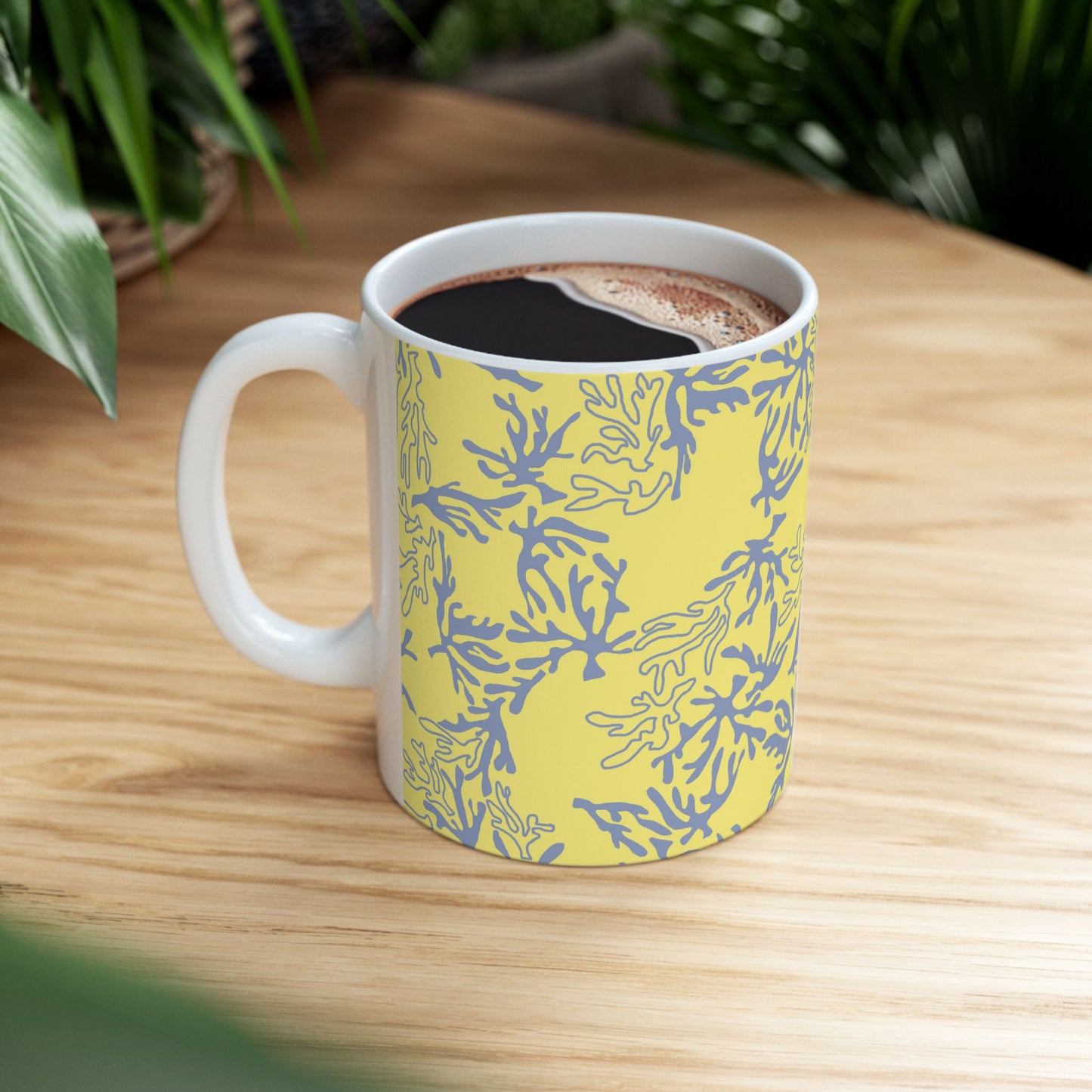 A white ceramic coffee mug with a yellow and blue coral pattern, filled with black coffee and sitting on a light wood table with green plants in the background.