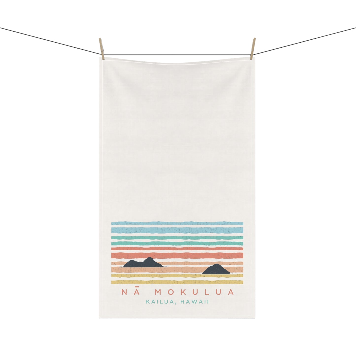 An off-white cotton tea towel hanging from a clothesline with wooden clips, featuring a retro-style sunset graphic with horizontal pastel stripes and the text 'NÁ MOKULUA KAILUA, HAWAII'.