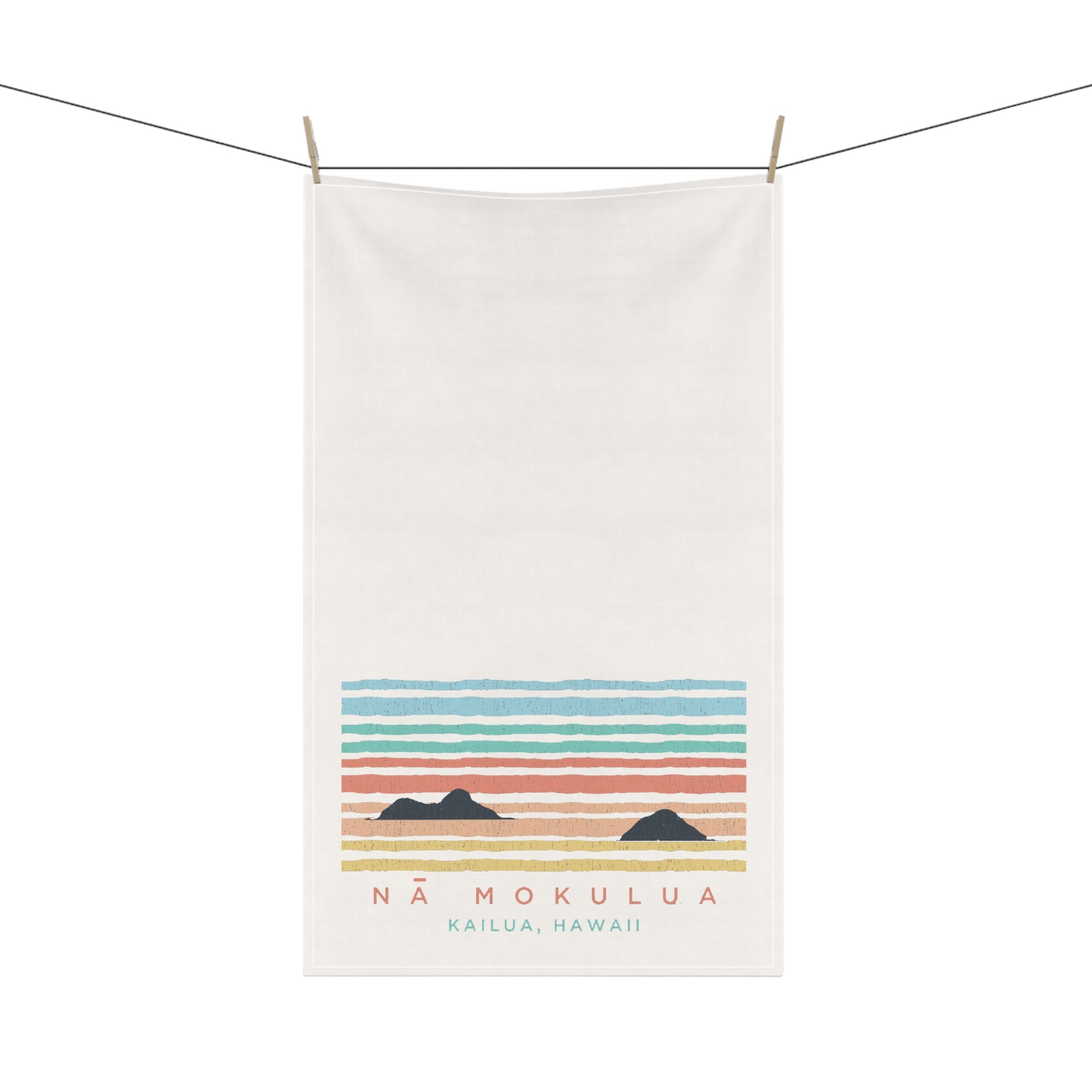 An off-white cotton tea towel hanging from a clothesline with wooden clips, featuring a retro-style sunset graphic with horizontal pastel stripes and the text 'NÁ MOKULUA KAILUA, HAWAII'.