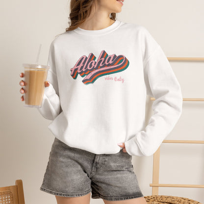 A woman wearing a white crewneck sweatshirt with a colorful retro-style 'Aloha Vibes Only' graphic, paired with gray denim shorts and holding an iced coffee.