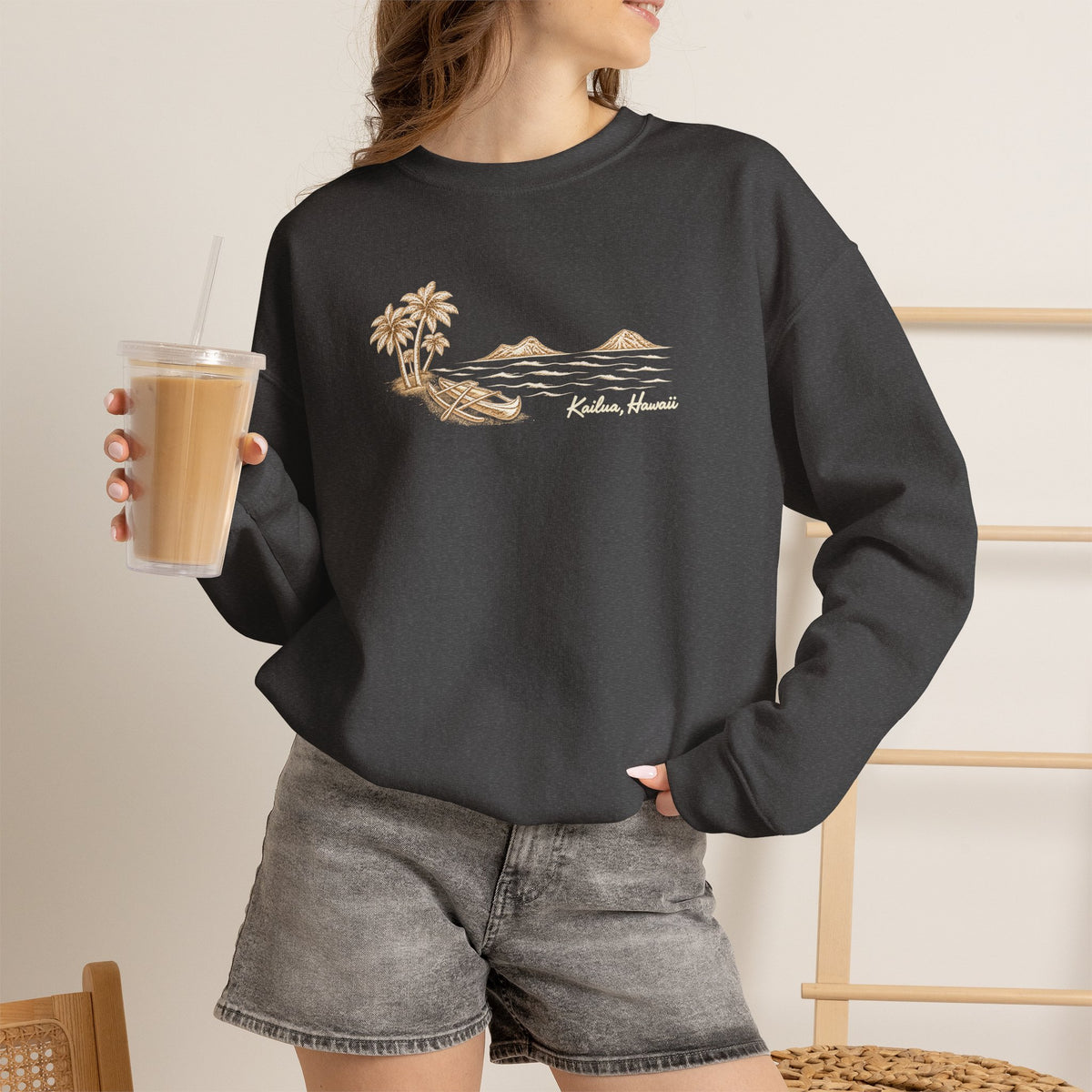 Woman standing holding an iced coffee wearing a Reverse hand-drawn Kailua canoe and Mokulua Islands unisex dark grey sweatshirt with ivory and tan artwork