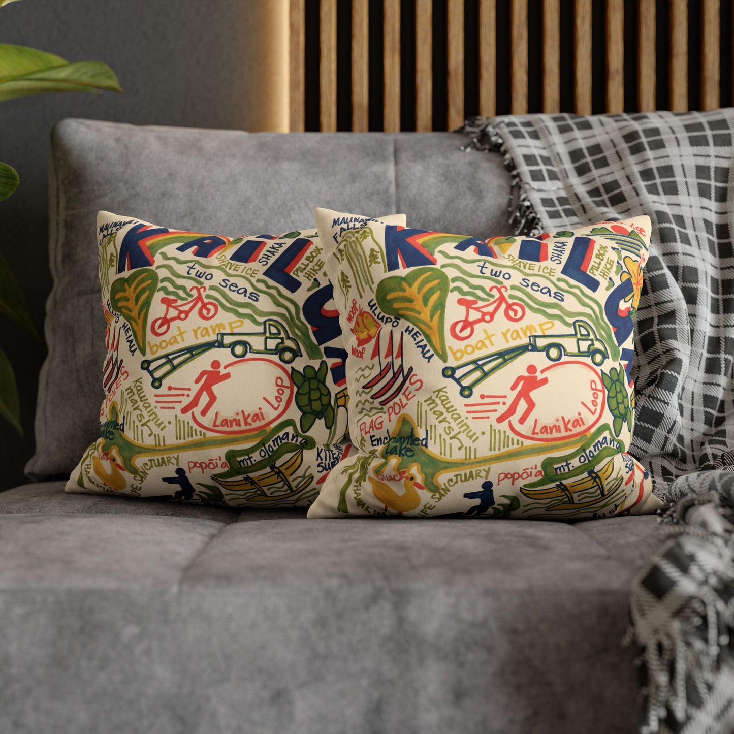 Two decorative throw pillows with a colorful vintage-style map and bicycle illustration pattern of Kailua, Oahu styled on a gray fabric sofa with a plaid throw blanket.