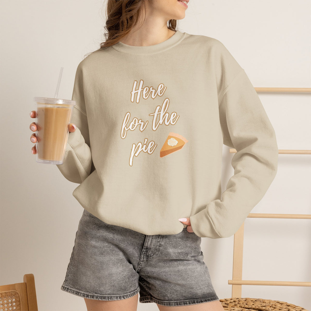 A woman wearing a sand crewneck sweatshirt with a graphic that reads 'Here for the pie' in brown script next to a slice of pie, paired with gray denim shorts and holding an iced coffee.