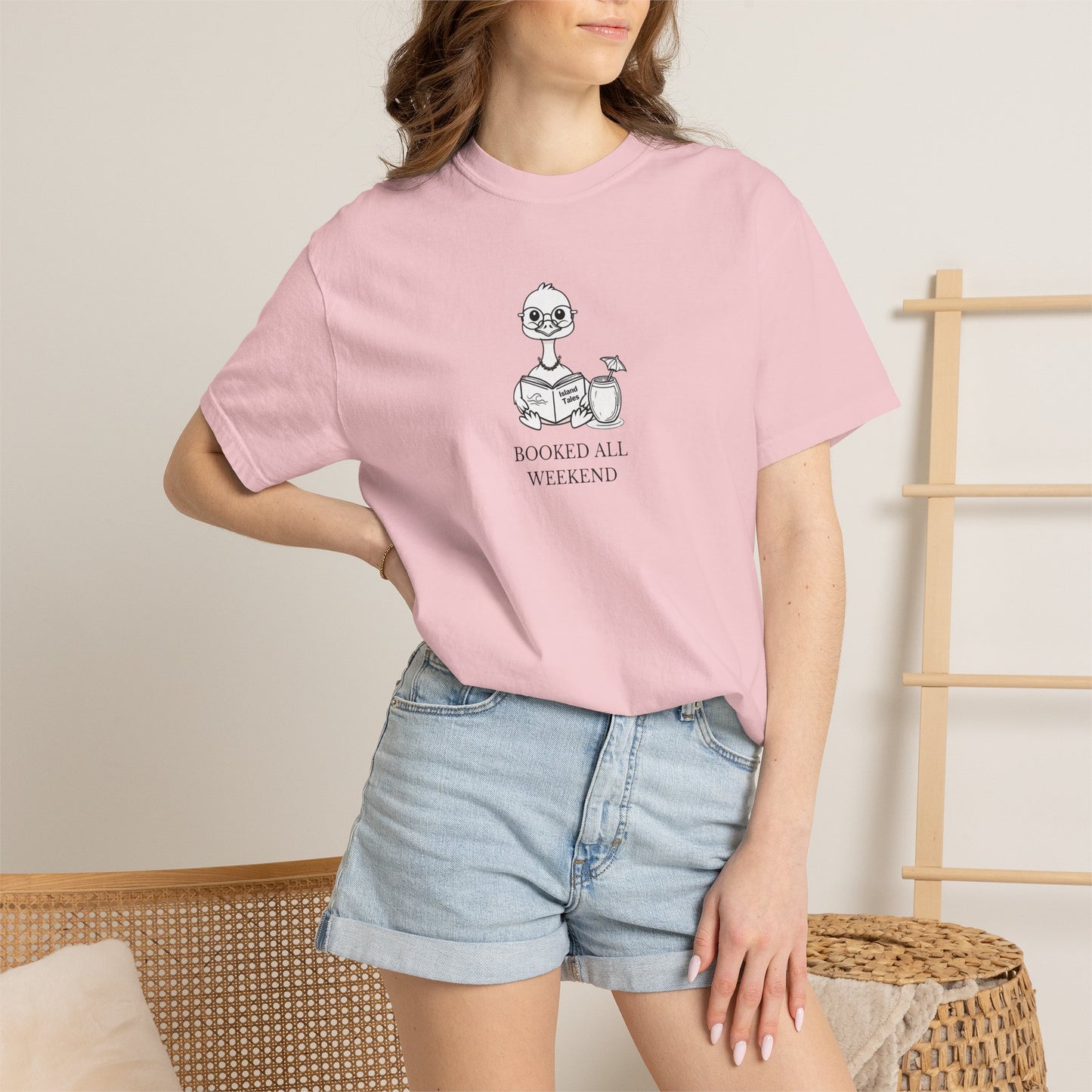 A woman standing wearing a pink comfort colors t-shirt with design of a nene goose reading a book and text 'booked all weekend' paired with denim shorts