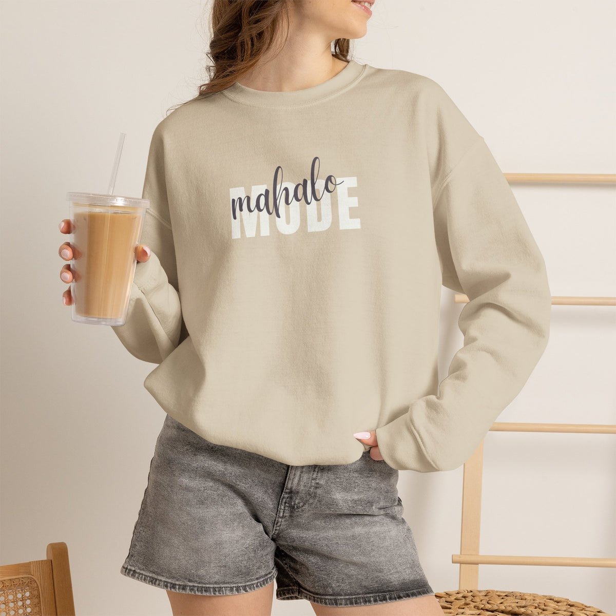 A woman wearing a sand crewneck sweatshirt with 'mahalo MODE' printed across the chest, featuring 'mahalo' in a dark gray script font and 'MODE' in a white block font, paired with washed black shorts and holding an iced coffee.