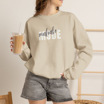 A woman wearing a sand crewneck sweatshirt with 'mahalo MODE' printed across the chest, featuring 'mahalo' in a dark gray script font and 'MODE' in a white block font, paired with washed black shorts and holding an iced coffee.