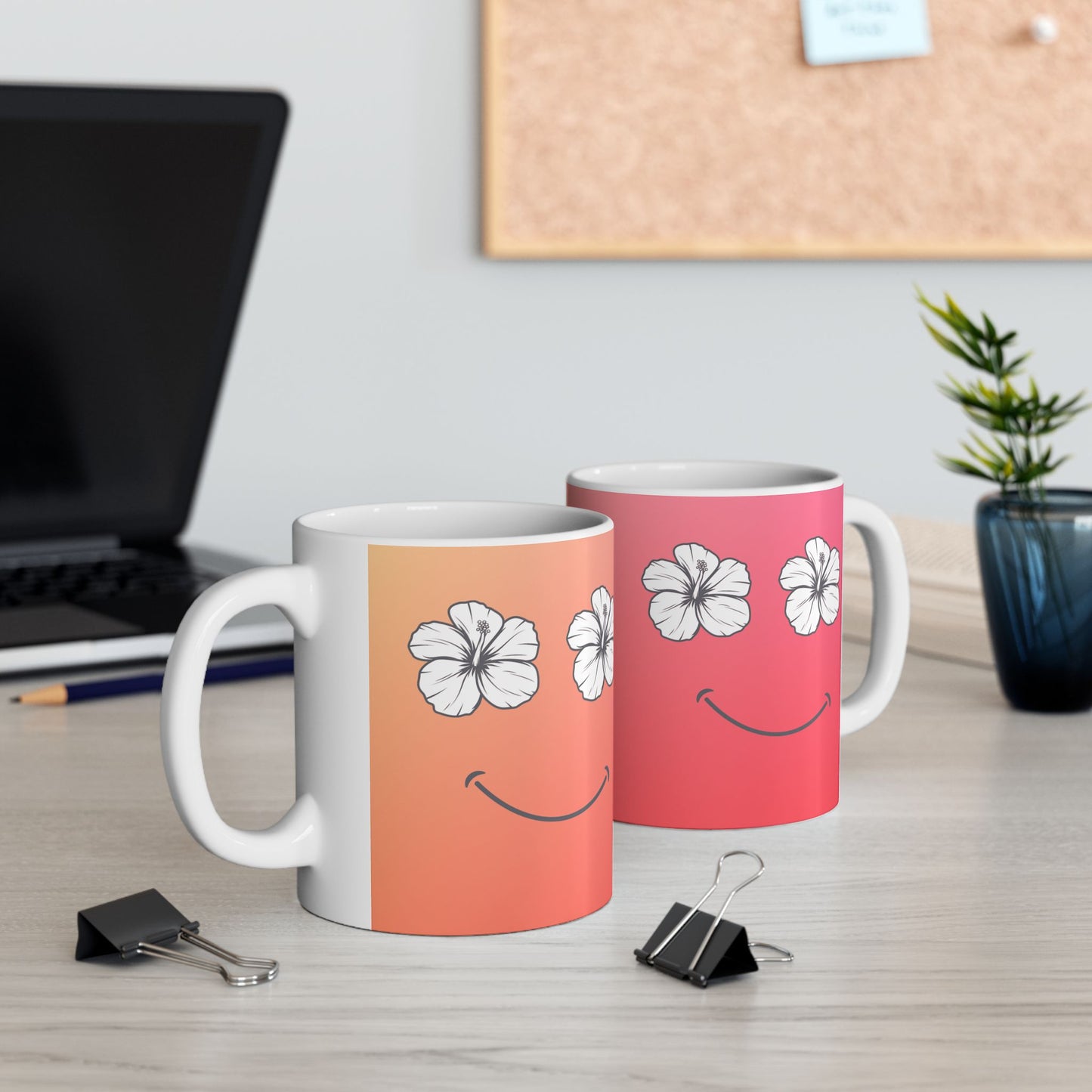 Two ceramic mugs with white handles and bases, decorated with a white hibiscus flower and smiley face design over a peach-to-pink gradient, displayed on a light wood desk with office props in the background.