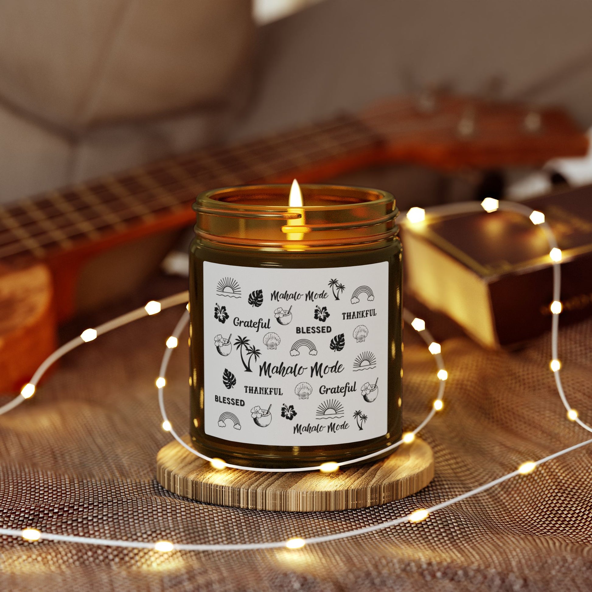 A dark amber glass jar candle with a gold metal screw-on lid and a white paper label featuring black hand-drawn style illustrations and the words 'Mahalo • Mode', 'Grateful', 'BLESSED', and 'THANKFUL'.on a wooden coaster, surrounded by string lights and a guitar in the background.