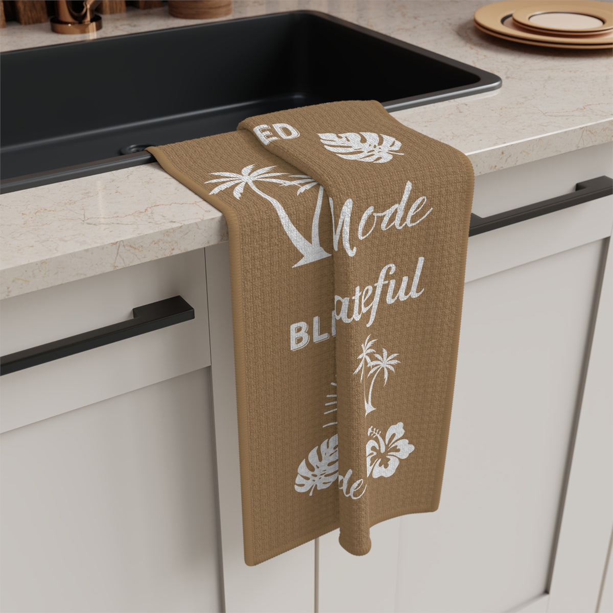 A tan kitchen towel with white tropical-themed illustrations and phrases including rainbows, palm trees, hibiscus flowers, seashells, and repeated text reading 'Grateful Mahalo Mode' and 'Blessed.' hanging off a kitchen counter