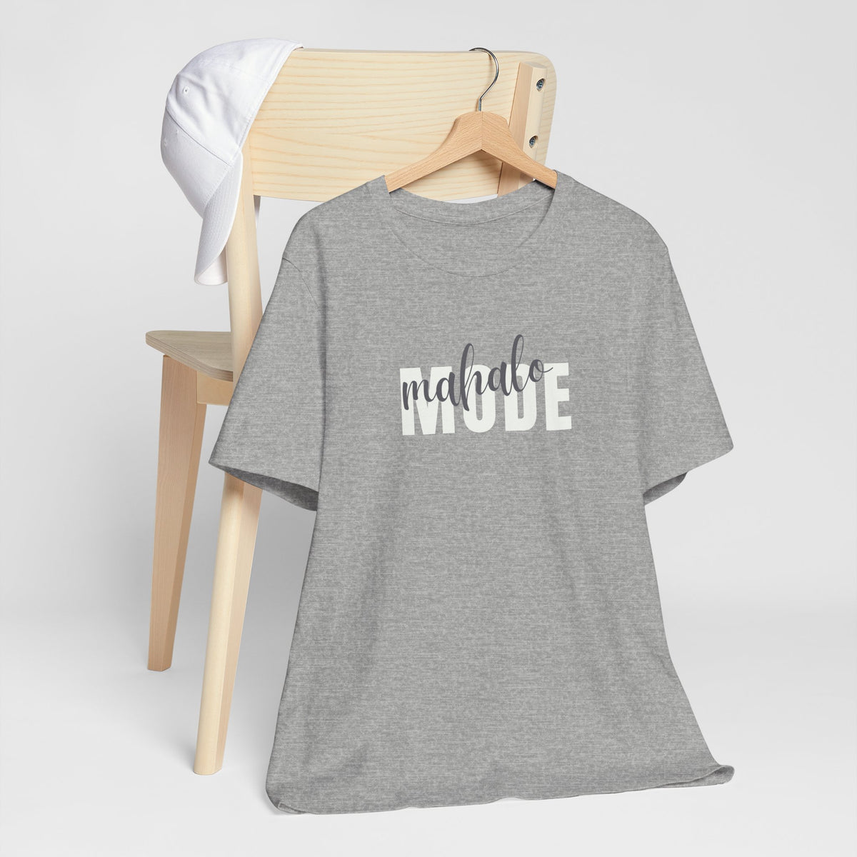 A heather gray short-sleeve t-shirt with 'mahalo MODE' printed across the chest, hanging on a wooden hanger in front of a light wood chair with a white towel draped over the back.